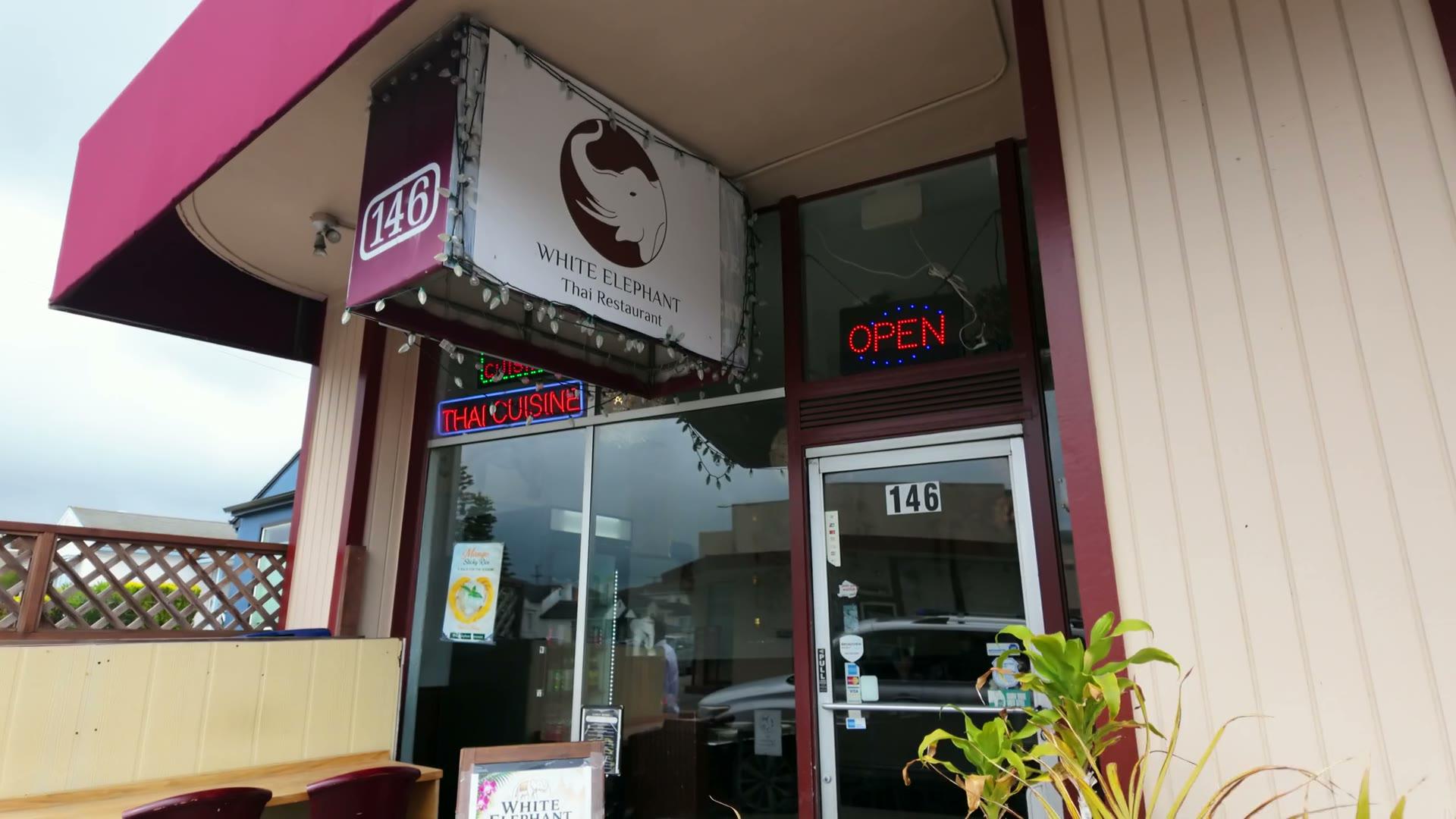The storefront at 146 Hazelwood Drive: the maroon awning, the White Elephant sign hung with fairy lights over the door, the OPEN sign lit in the window and a neon THAI CUISINE sign beside it.