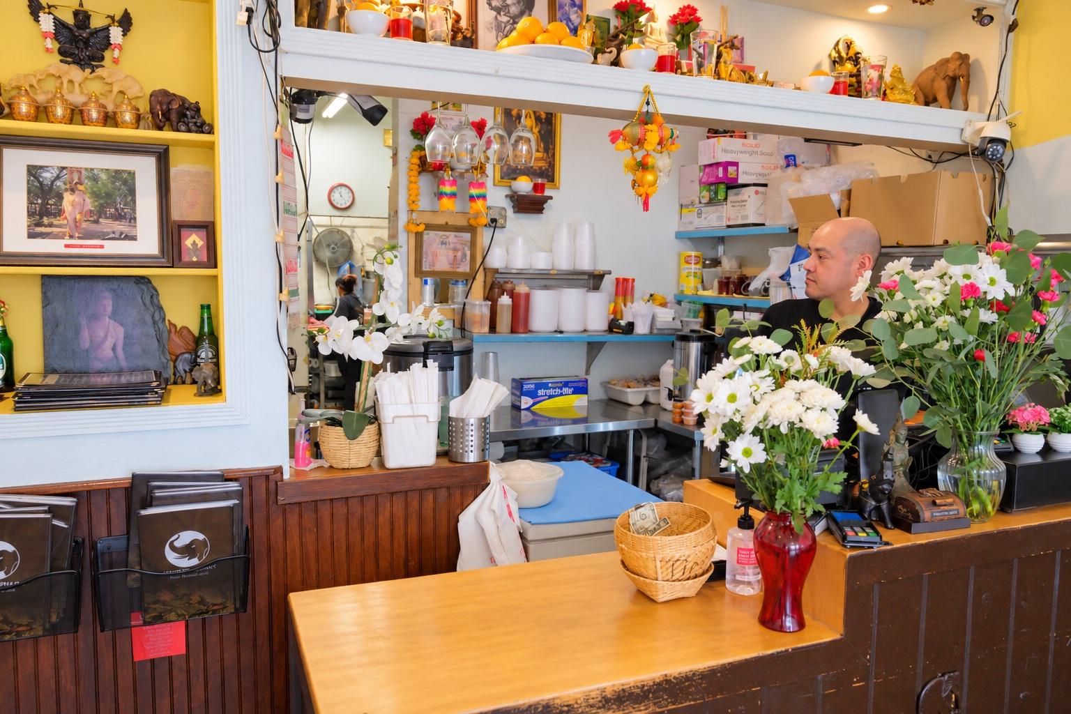 The front counter with fresh flowers and a card reader, shelves of figures, fruit and framed pictures above, a staff member behind it, and the kitchen pass beyond.