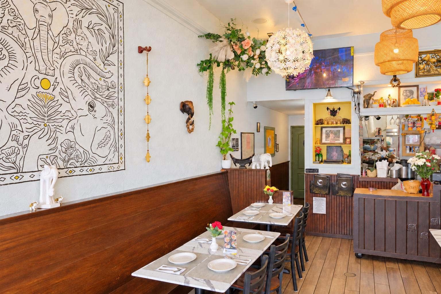 The whole dining room from the door: the elephant mural on the left wall above the wood wainscot, marble tables set with plates, a flowered chandelier, and the front counter at the back.