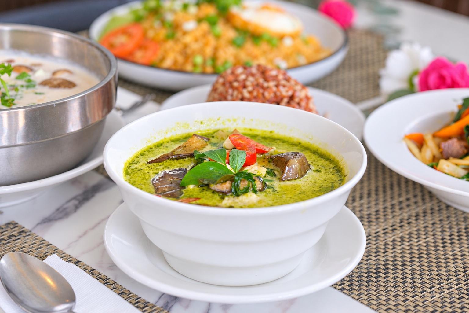 A white bowl of green curry with aubergine, red chilli and basil in the foreground; a steel bowl of coconut soup, a plate of fried rice with a fried egg, and brown rice behind it.